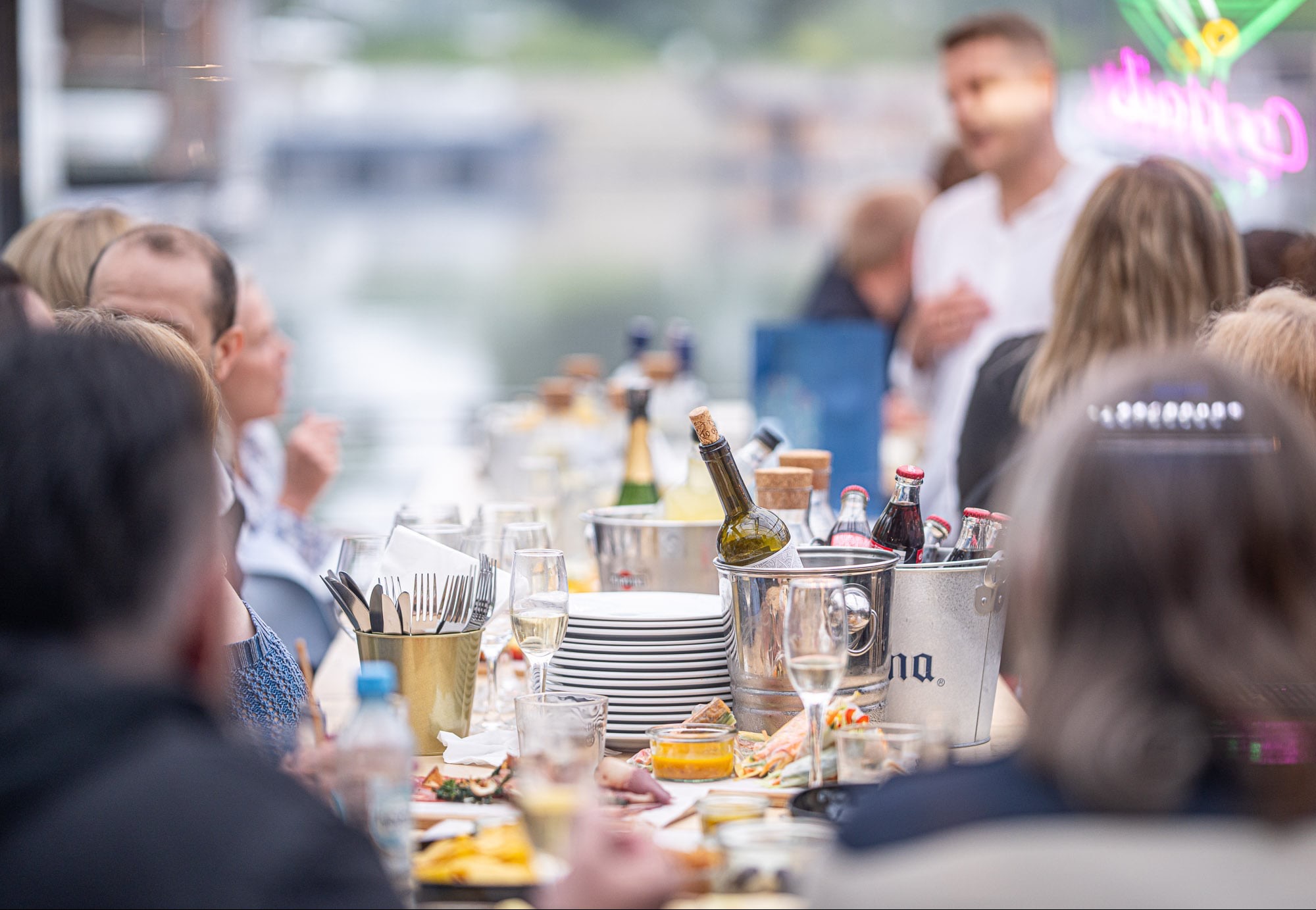 Uroczysty stół z napojami w wiaderkach, zastawą i jedzeniem, otoczony przez gości, z neonowym napisem w tle, sugerujący eleganckie przyjęcie.
