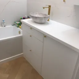 Nowoczesna łazienka z białą szafką na wymiar, marmurową umywalką i złotą armaturą. Parkiet w jodełkę. Minimalistyczny design z akcentami luksusu.