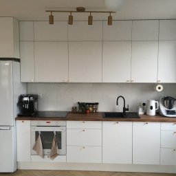 Peleda - Nowoczesna, jasna kuchnia z białymi szafkami i drewnianym blatem. Złote akcenty oświetlenia i uchwytów dodają elegancji. Widoczny sprzęt AGD i akcesoria kuchenne.