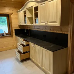 Jasna kuchnia w drewnianym domu: kremowe szafki z frezowanymi frontami, czarny blat i złota bateria. Otwarta szuflada ukazuje wnętrze. Widok na okno i drewniane ściany.