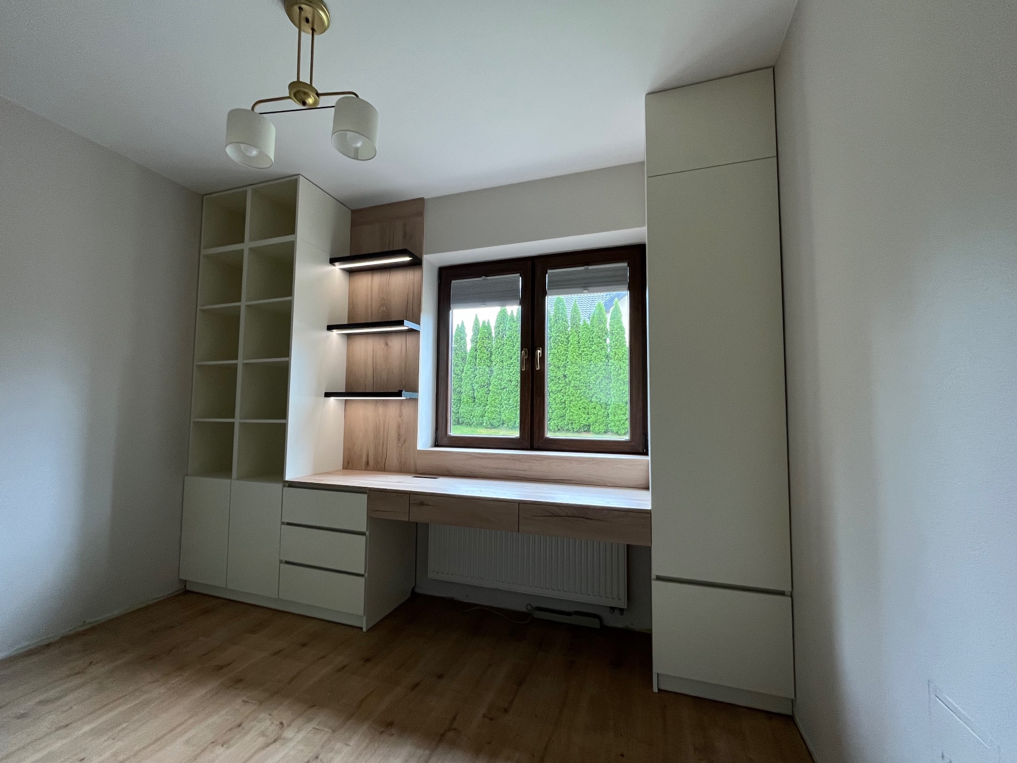 Nowoczesny pokój z zabudową meblową: białe szafy z półkami i biurko z drewnianym blatem pod oknem, oświetlenie punktowe, drewniana podłoga. Minimalistyczny design.