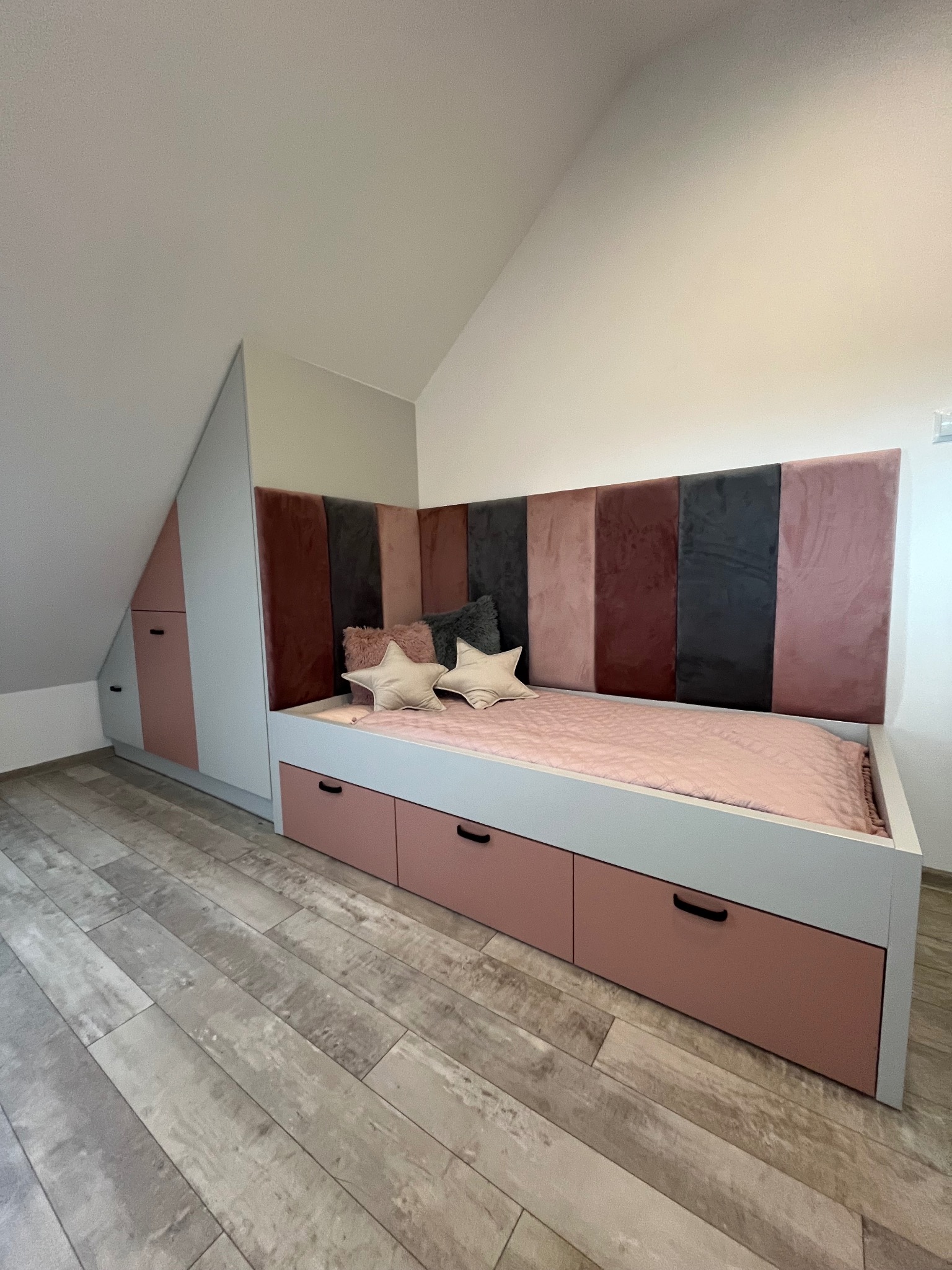 Pokój dziecięcy na poddaszu: zabudowa szafy wnękowej z łóżkiem z szufladami, tapicerowany zagłówek w odcieniach różu i szarości, poduszki w kształcie gwiazd.