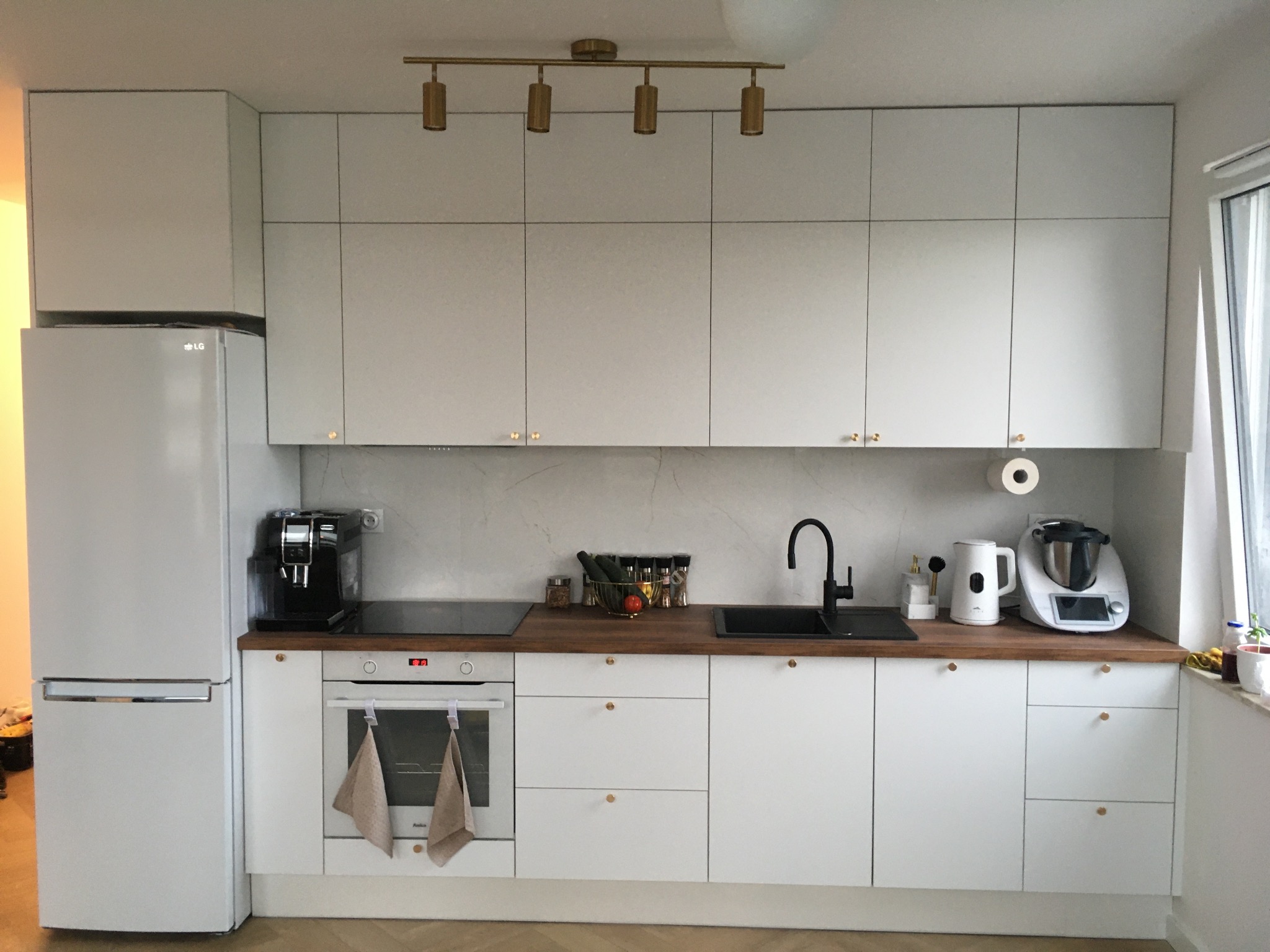 Nowoczesna, jasna kuchnia z białymi szafkami i drewnianym blatem. Złote akcenty oświetlenia i uchwytów dodają elegancji. Widoczny sprzęt AGD i akcesoria kuchenne.