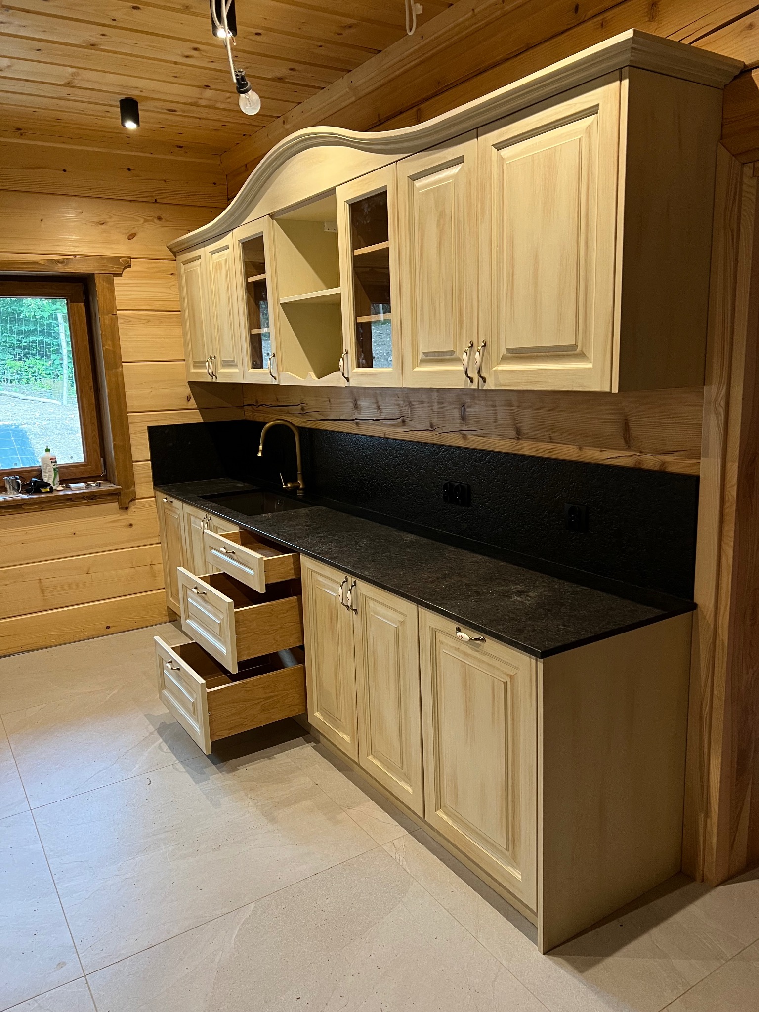 Jasna kuchnia w drewnianym domu: kremowe szafki z frezowanymi frontami, czarny blat i złota bateria. Otwarta szuflada ukazuje wnętrze. Widok na okno i drewniane ściany.