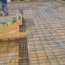 Zbrojenie stropu budynku jednorodzinnego, widoczne drewniane szalunki i siatka z prętów zbrojeniowych na tle zabudowy mieszkalnej w pochmurny dzień.