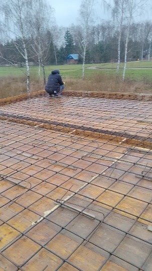 Zbrojenie fundamentu z siatki stalowej na deskowaniu, w tle pracownik w kurtce, drzewa i budynek. Prace budowlane w pochmurny dzień.