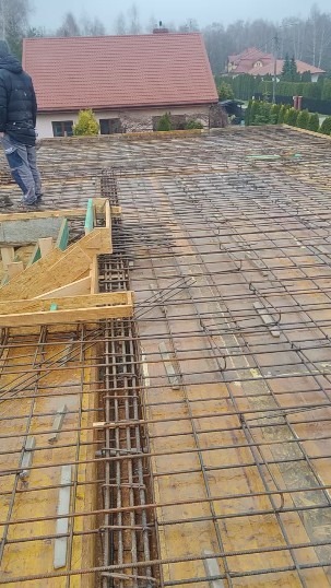 Zbrojenie stropu budynku jednorodzinnego, widoczne drewniane szalunki i siatka z prętów zbrojeniowych na tle zabudowy mieszkalnej w pochmurny dzień.