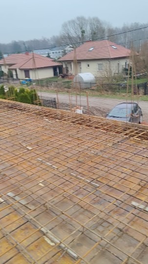 Zbrojenie stropu domu jednorodzinnego z widocznymi prętami i deskowaniem, w tle zabudowa mieszkalna i samochód. Ujęcie z góry, pochmurny dzień.