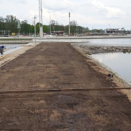 Łukasz Jankowski Tu Mi Kop - Niwelacja terenu podłoża na nabrzeżu z widoczną ziemią, częściowo drewnianym pomostem i wózkiem budowlanym w tle, w pobliżu zbiornika wodnego.