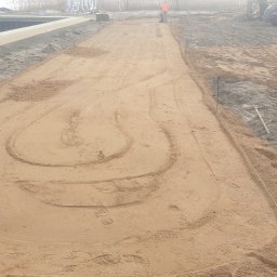 Łukasz Jankowski Tu Mi Kop - Niwelacja terenu piaskiem, widoczne ślady opon i stopy, wytyczone granice sznurkiem. W tle osoba obsługująca maszynę i zaparkowany samochód osobowy.
