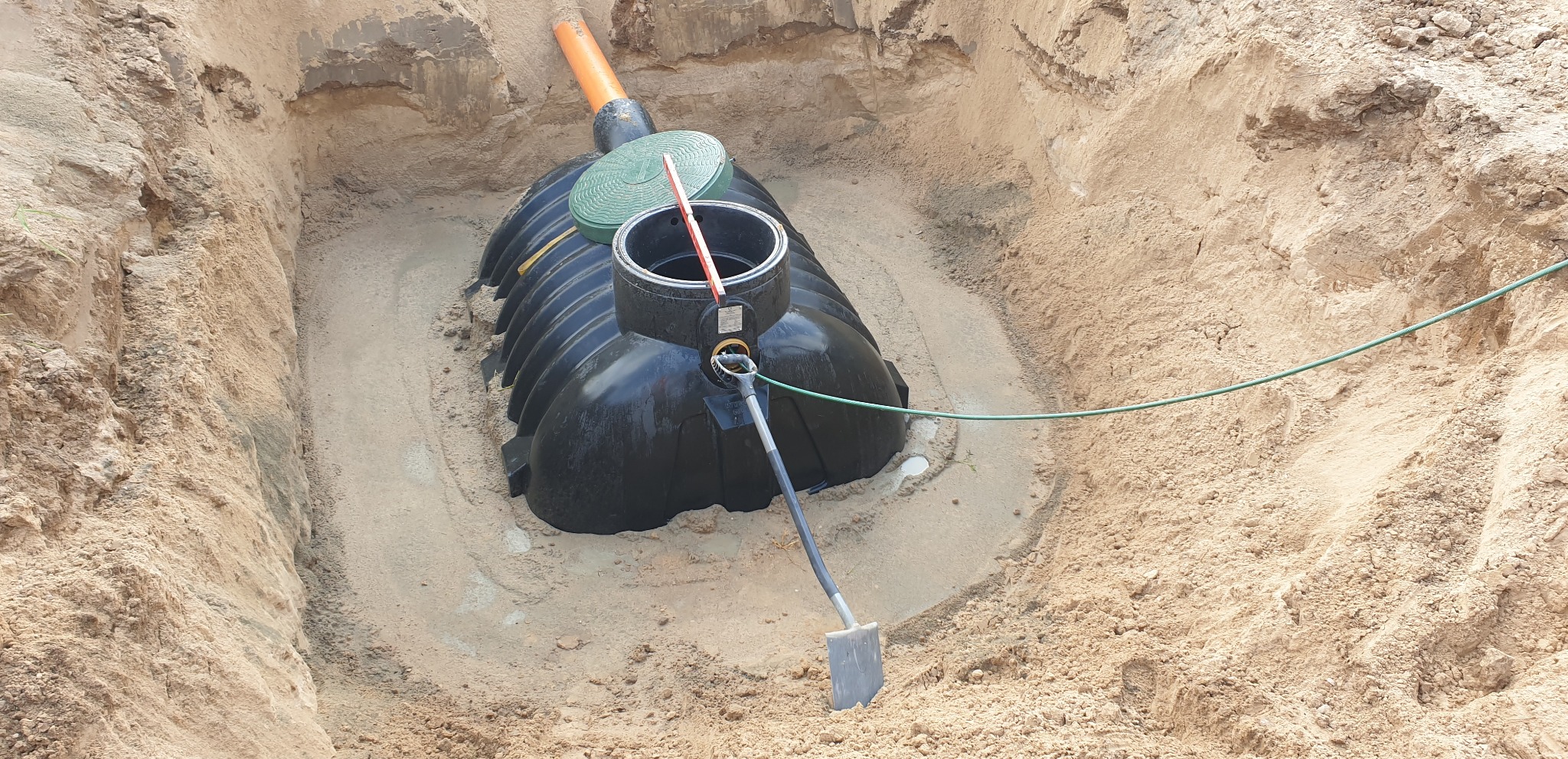 Czarny zbiornik na szambo umieszczony w wykopie z piasku, widoczna łopata i poziomica, pomarańczowa rura i zielona pokrywa.