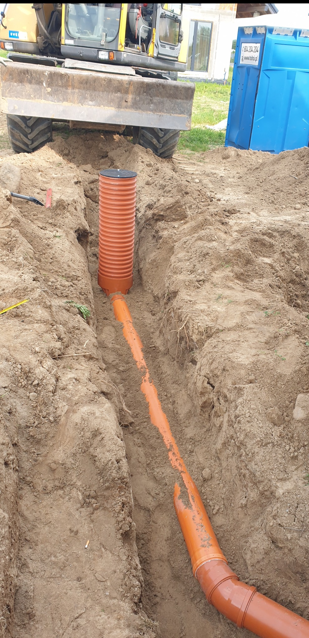 Pomarańczowe rury drenażowe ułożone w wykopie ziemnym, z widocznym zbiornikiem drenażowym i koparką w tle. Instalacja drenażu w trakcie realizacji.