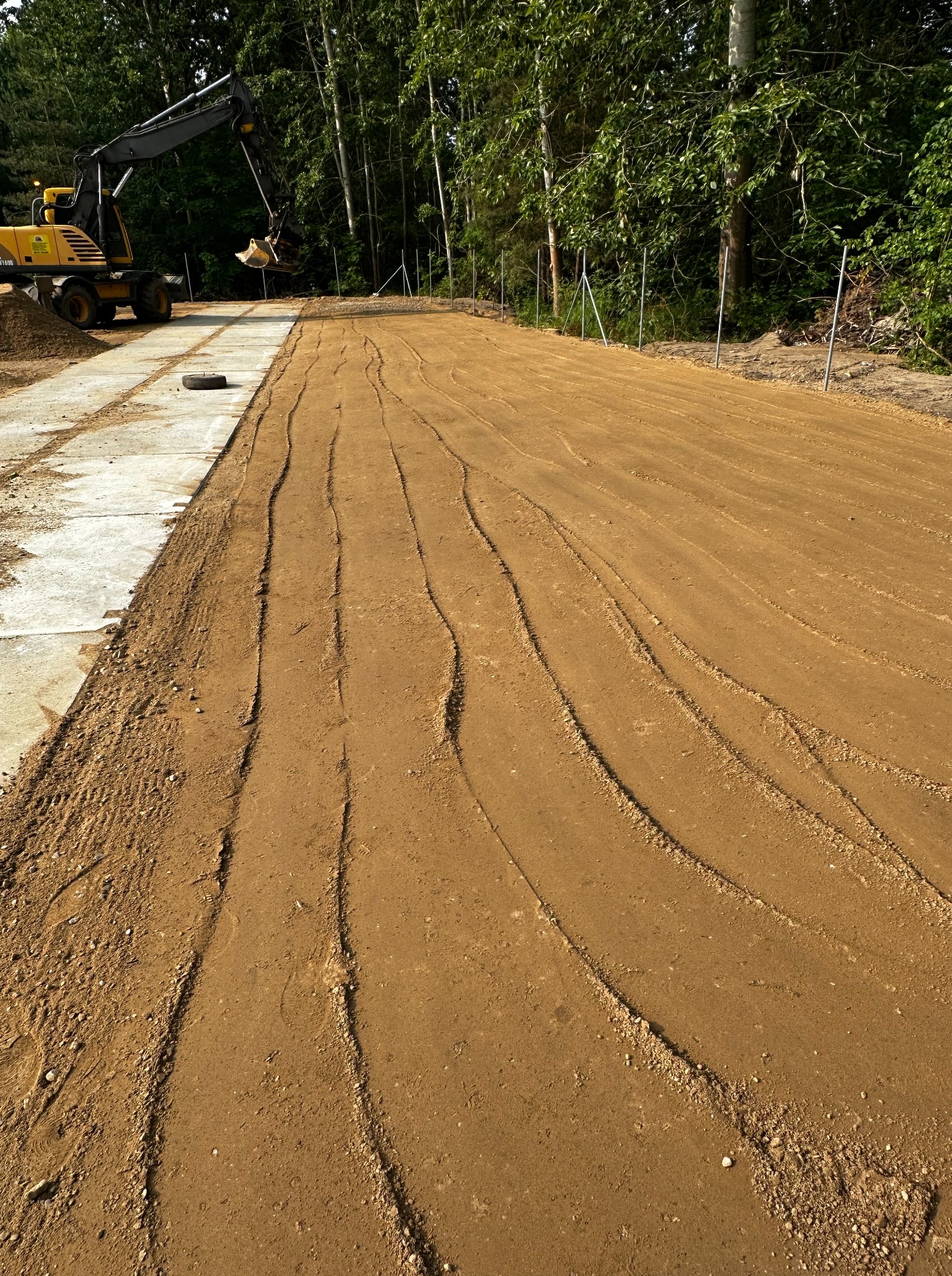 Żółta koparka wyrównuje teren pod budowę ogrodzenia, widoczne ślady opon na piasku i wbite słupki ogrodzeniowe w tle. Prace ziemne w toku, widok z góry.