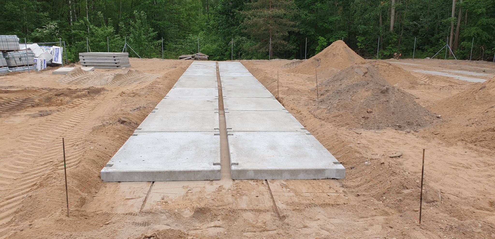 Ułożone betonowe płyty na piasku, tworzące tymczasową drogę na placu budowy. Widoczne ślady opon, hałdy piachu i drzewa w tle. Płyty z charakterystycznymi wgłębieniami na rogach.