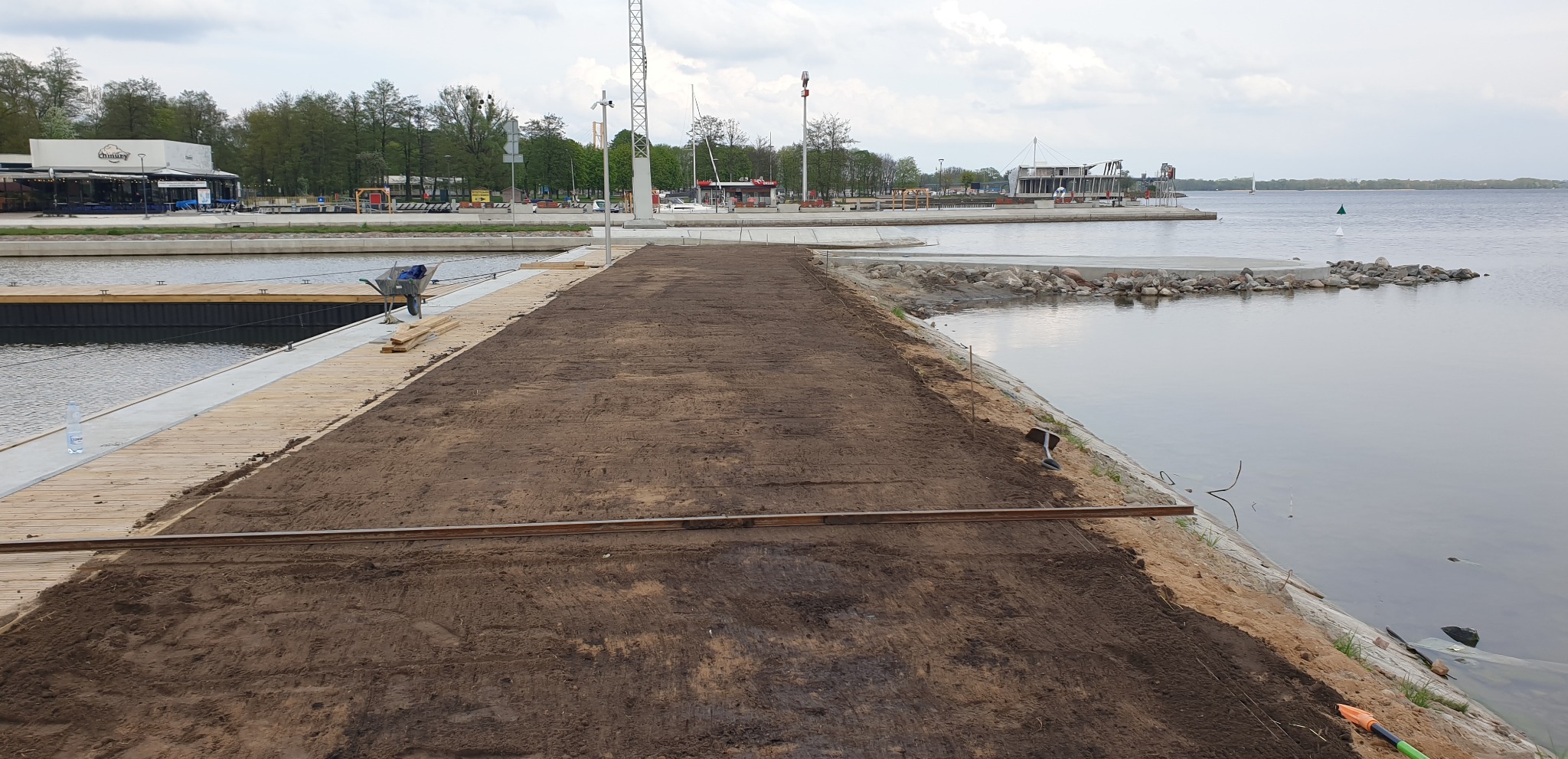 Niwelacja terenu podłoża na nabrzeżu z widoczną ziemią, częściowo drewnianym pomostem i wózkiem budowlanym w tle, w pobliżu zbiornika wodnego.