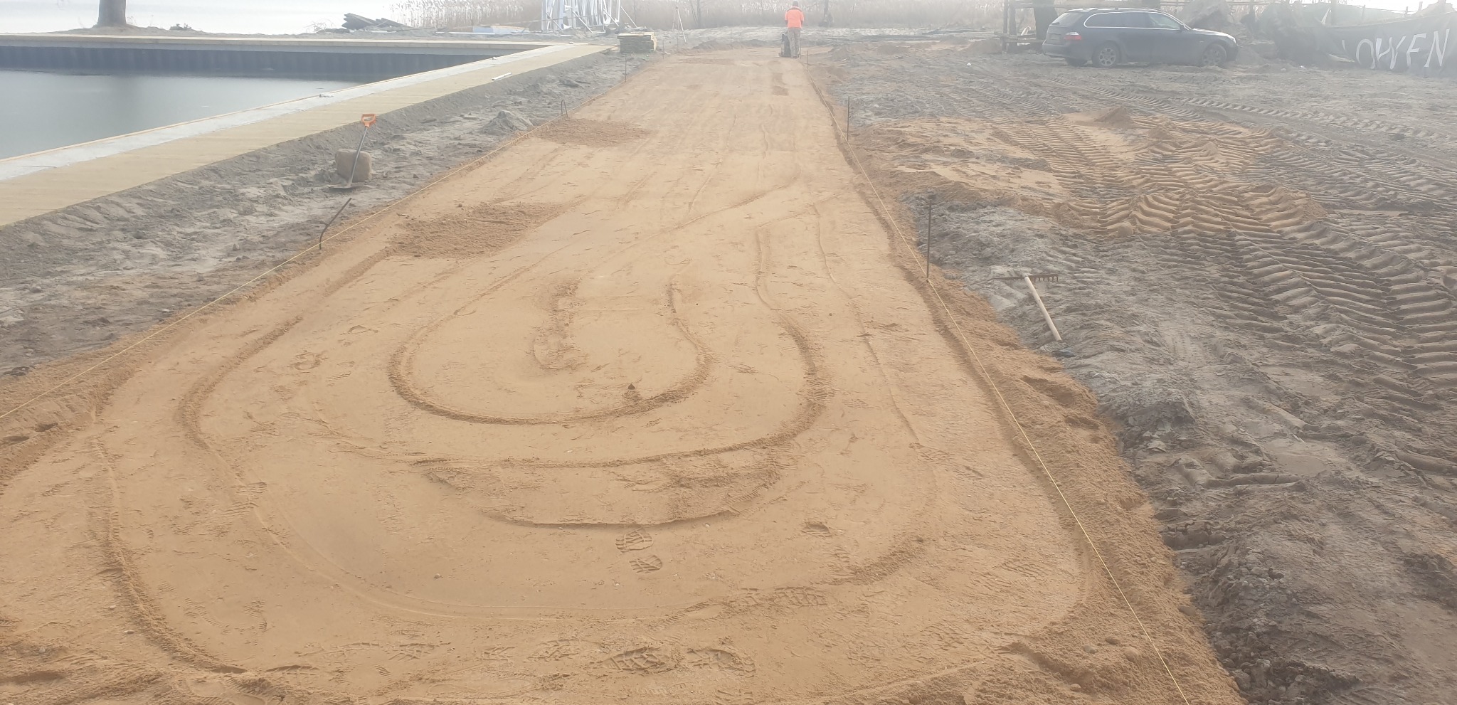 Niwelacja terenu piaskiem, widoczne ślady opon i stopy, wytyczone granice sznurkiem. W tle osoba obsługująca maszynę i zaparkowany samochód osobowy.