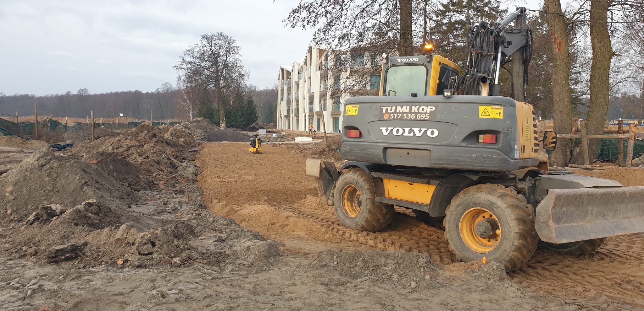 Szaro-żółta koparka Volvo na placu budowy, w tle budynek mieszkalny i drzewa. Widać wyrównany teren przygotowany pod dalsze prace. Na koparce logo firmy Tumi Kop.
