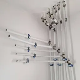 Białe rury instalacji wodnej zamocowane do ściany i sufitu za pomocą metalowych uchwytów z niebieskimi elementami. Widoczne liczne złączki i kolanka.