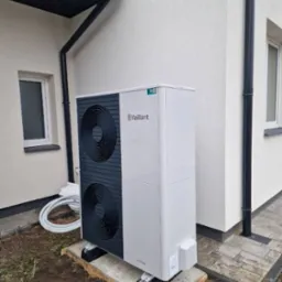 Nowoczesna pompa ciepła Vaillant zamontowana przy budynku z jasną elewacją. Widoczne czarne rury spustowe i okno. Estetyczny montaż na drewnianej podstawie.