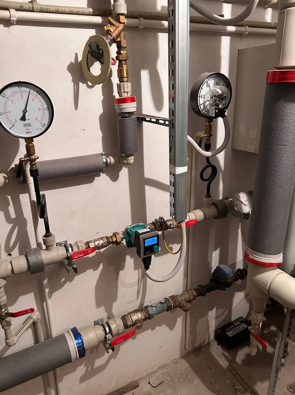 Węzeł cieplny z manometrami, zaworami i pompą obiegową Wilo, rury zaizolowane szarą pianką. Widoczne przyłącza i metalowa szyna montażowa.