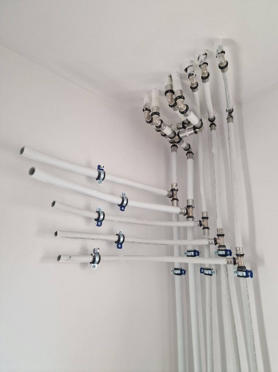 Białe rury instalacji wodnej zamocowane do ściany i sufitu za pomocą metalowych uchwytów z niebieskimi elementami. Widoczne liczne złączki i kolanka.