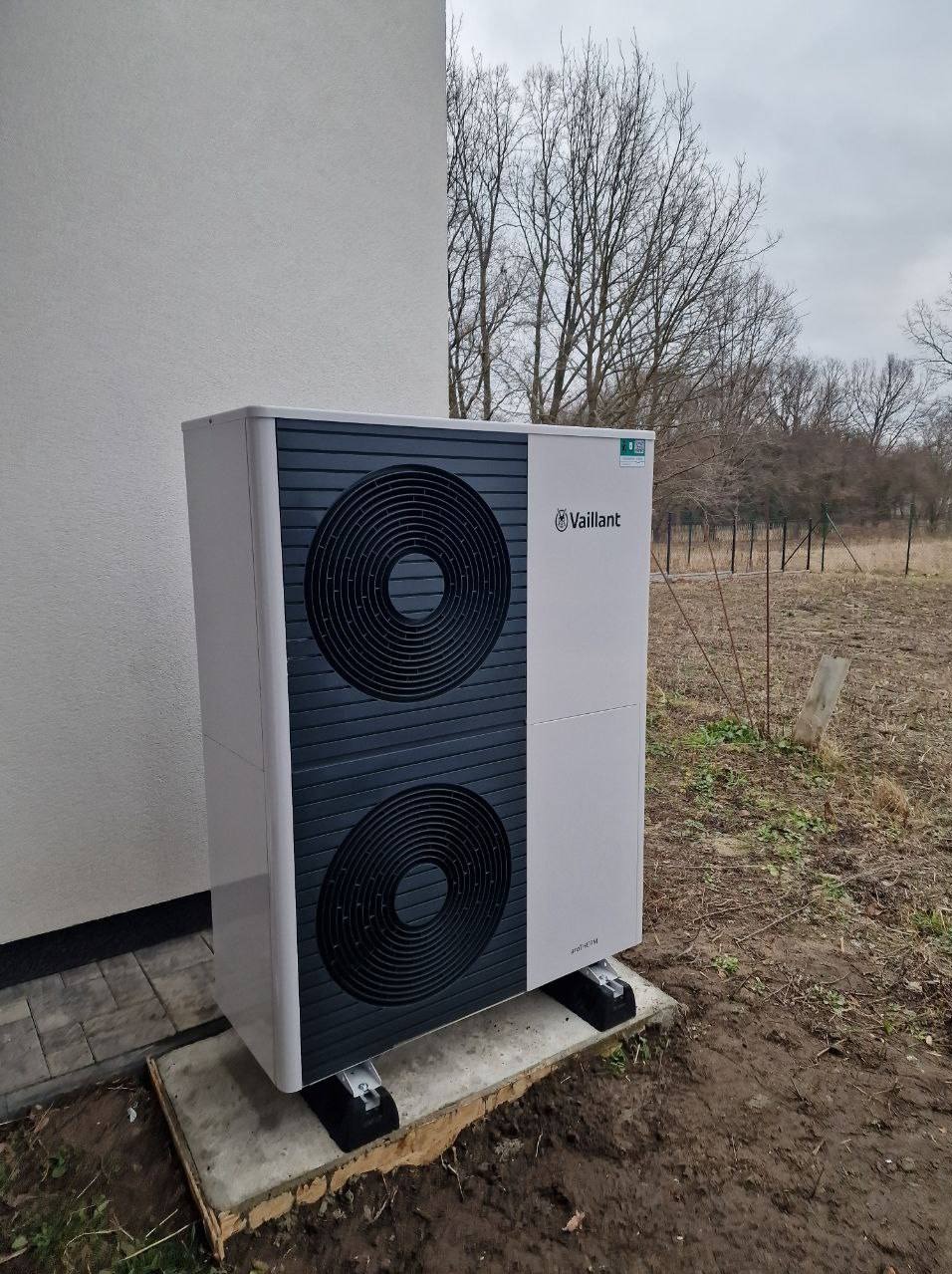 Zewnętrzna pompa ciepła Vaillant aroTHERM split na betonowej podstawie, zamontowana przy ścianie budynku. Widoczne drzewa w tle.