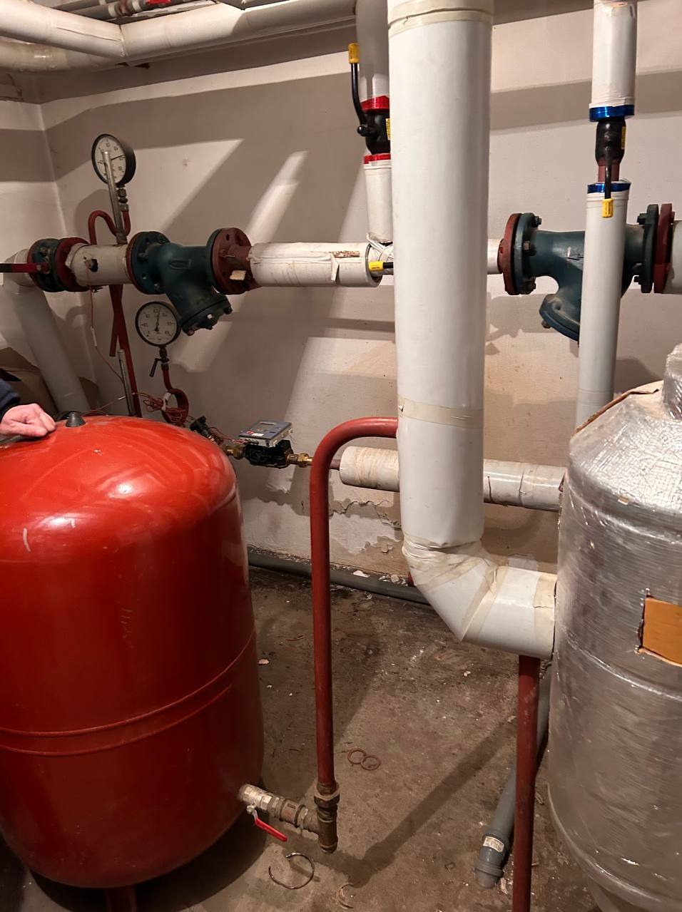 Czerwony zbiornik wyrównawczy obok izolowanych rur i zaworów w kotłowni. Manometry na ścianie. Instalacja z bliska, widoczne łączenia i elementy armatury.