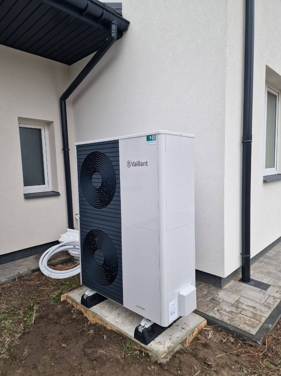 Nowoczesna pompa ciepła Vaillant zamontowana przy budynku z jasną elewacją. Widoczne czarne rury spustowe i okno. Estetyczny montaż na drewnianej podstawie.
