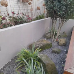 Kaskadowy ogród z roślinami w podwyższonych rabatach, kamieniami i szarym żwirem. Drewniany element architektury ogrodowej widoczny z boku.