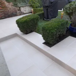 Jasne płytki tarasowe z dekoracyjnymi schodkami i geometrycznie przyciętymi krzewami bukszpanu, tworzące nowoczesną aranżację ogrodu przydomowego.