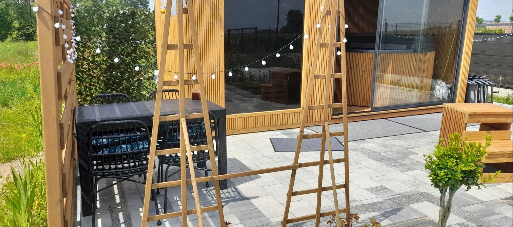 Wykończony taras z kostki brukowej z rattanowymi meblami ogrodowymi i drewnianą pergolą, oświetlony girlandami, obok nowoczesnego domku z jacuzzi.