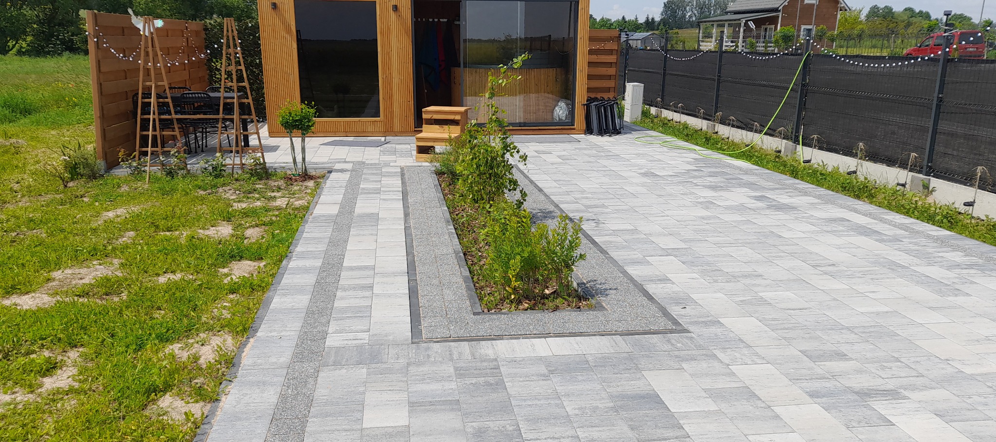 Szara kostka brukowa ułożona w geometryczny wzór, z wyznaczonym miejscem na rośliny, otoczona nowoczesnym domkiem i czarnym ogrodzeniem.