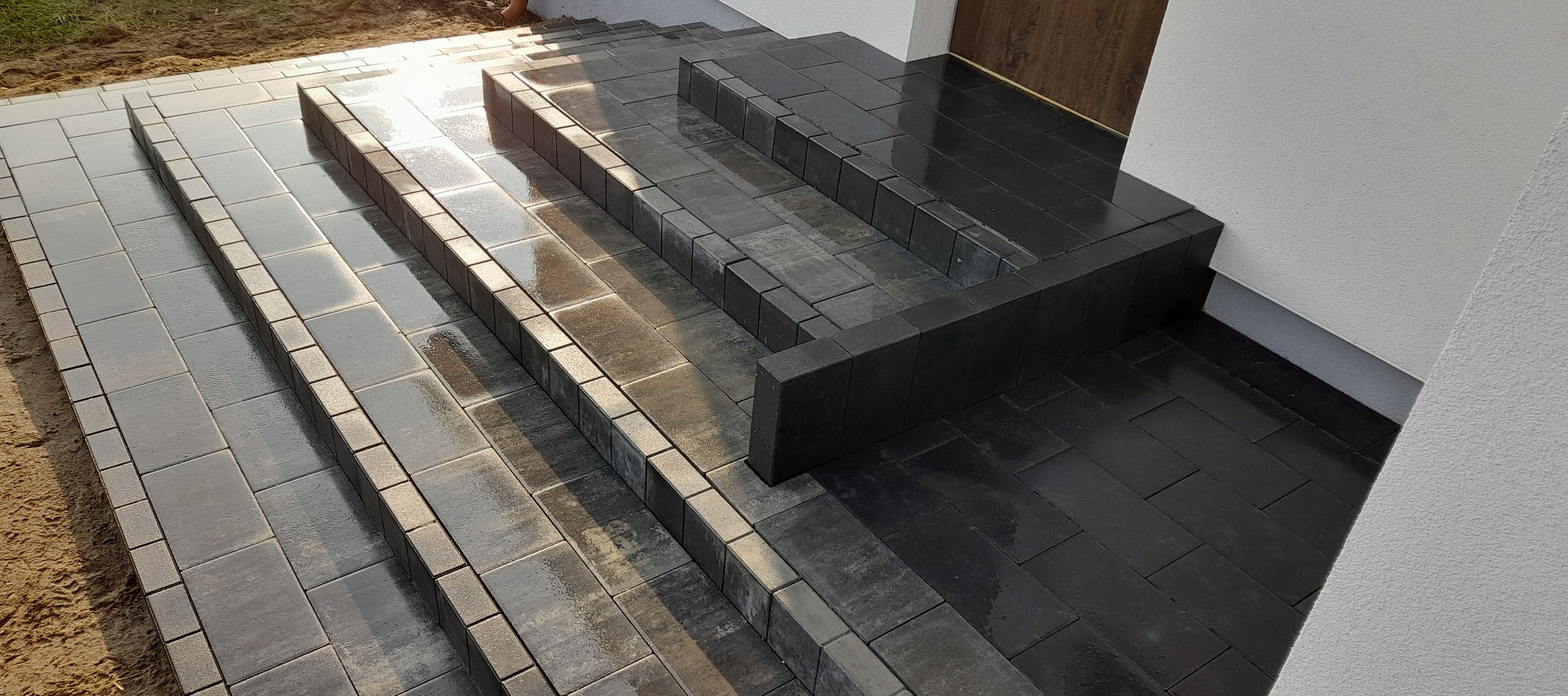 Schody wejściowe wyłożone ciemną kostką brukową, z obrzeżami w jaśniejszym odcieniu. Widoczna faktura kostki i delikatne odbicia światła. Estetyczne wykończenie.