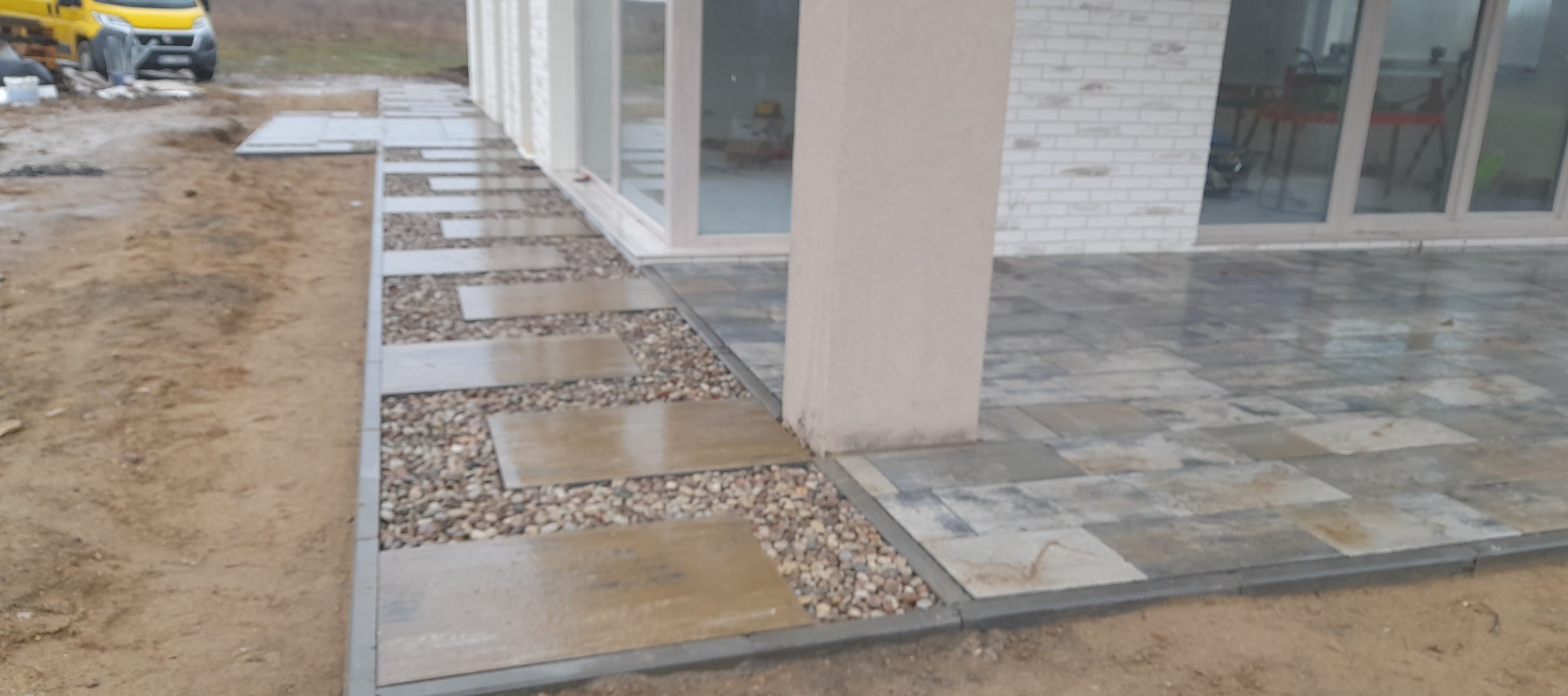 Ułożona ścieżka z płyt i kamieni ozdobnych wzdłuż domu z wybrukowanym tarasem. Widoczny fragment niezabudowanego terenu i zaparkowany samochód dostawczy.