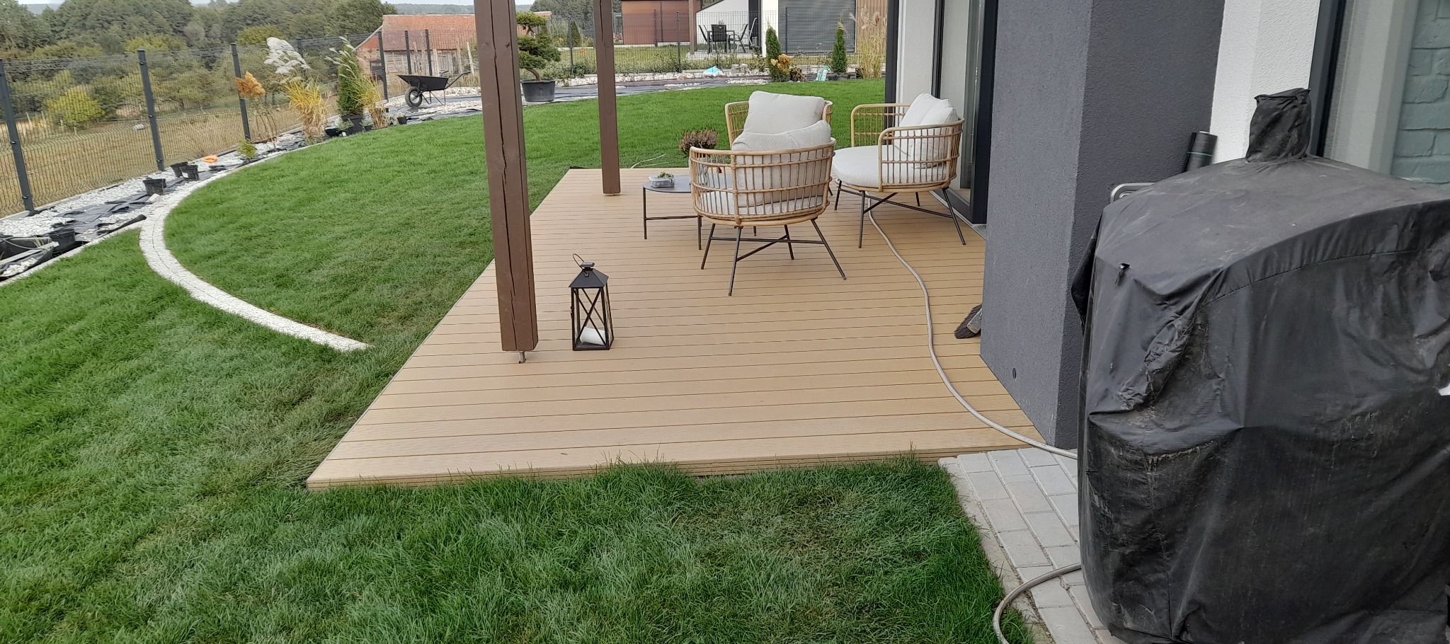 Drewniany taras z meblami ogrodowymi i lampionem, otoczony świeżym trawnikiem i ozdobnymi nasadzeniami. Widoczne elementy wykończeniowe i dbałość o detale.