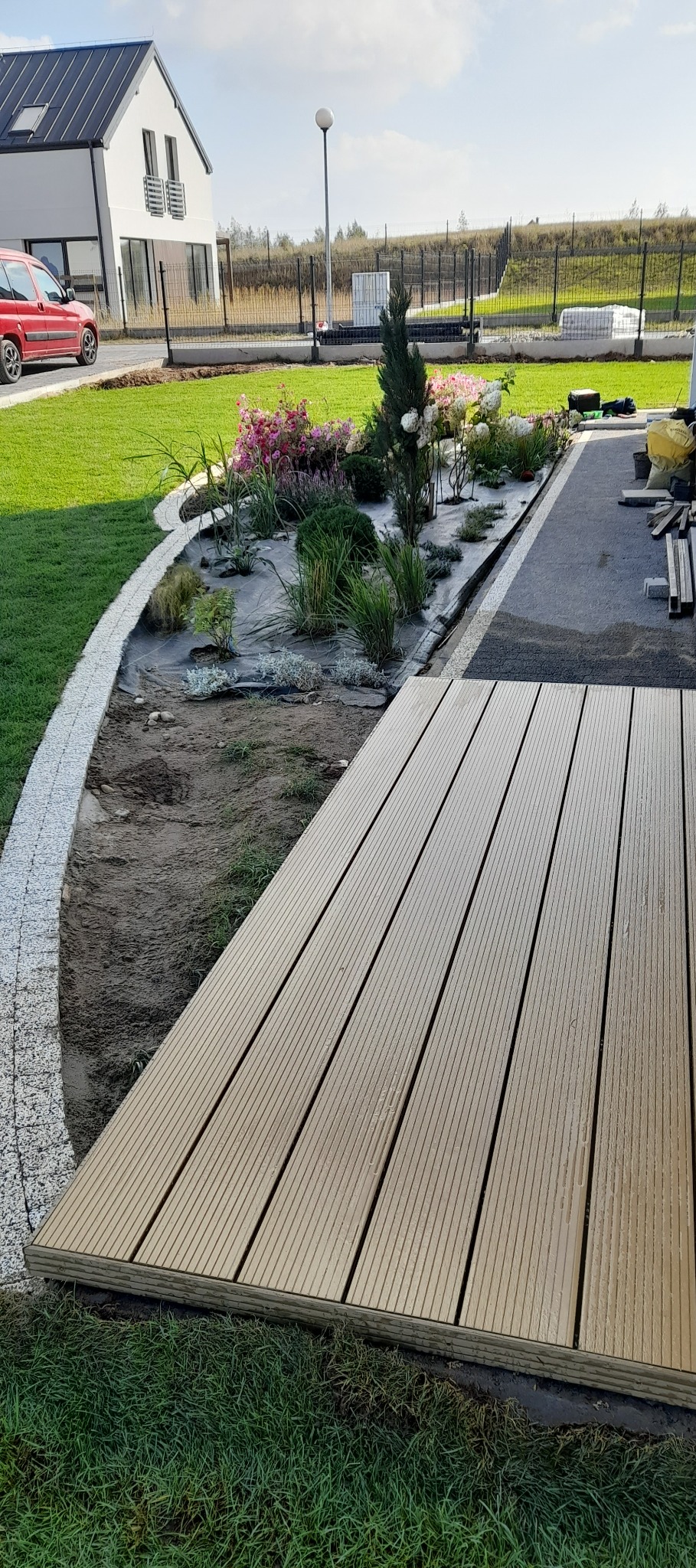 Nowoczesny ogród w trakcie realizacji: drewniany taras, rabata z roślinami i kostką granitową, dom w tle.