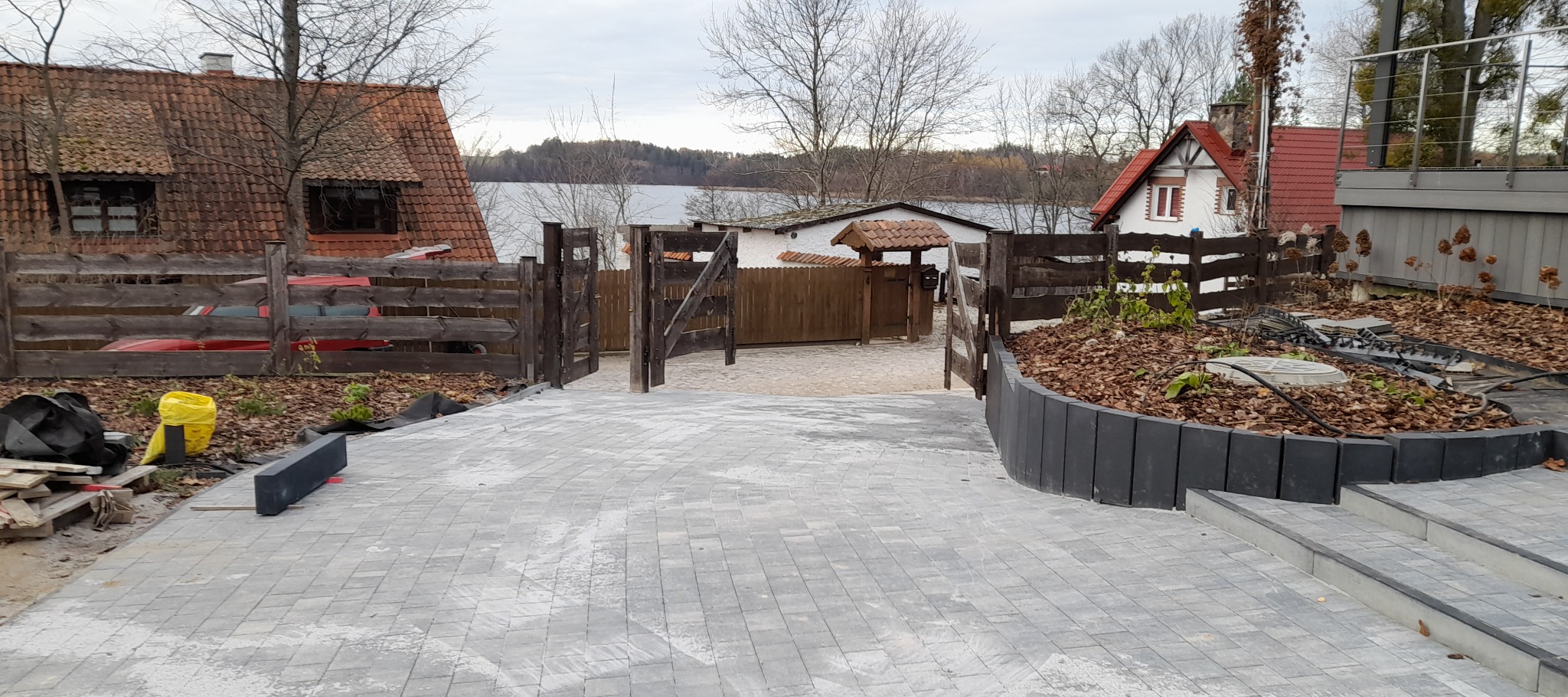 Nowa, szara kostka brukowa prowadząca do drewnianej bramy i zadbanego ogrodu z widokiem na jezioro w tle. Częściowo widoczny dom z czerwonym dachem.
