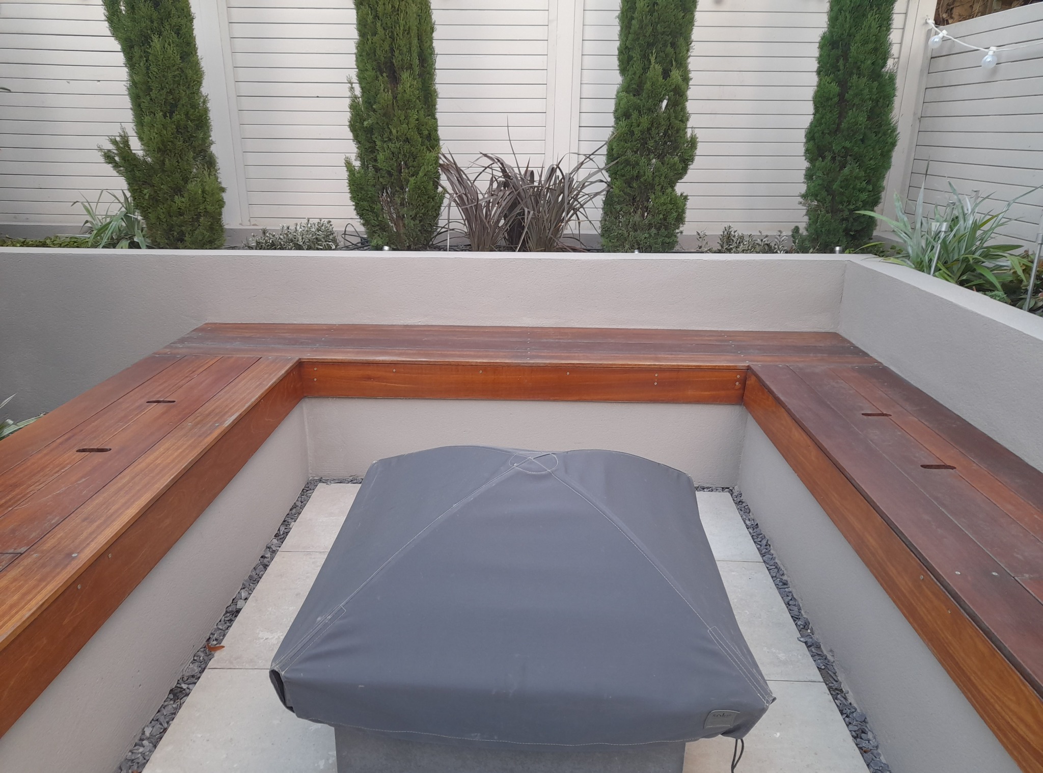 Nowoczesny ogród z wnęką na palenisko, drewnianymi ławkami i wysokimi, smukłymi drzewami w tle, tworzącymi intymną przestrzeń relaksu.