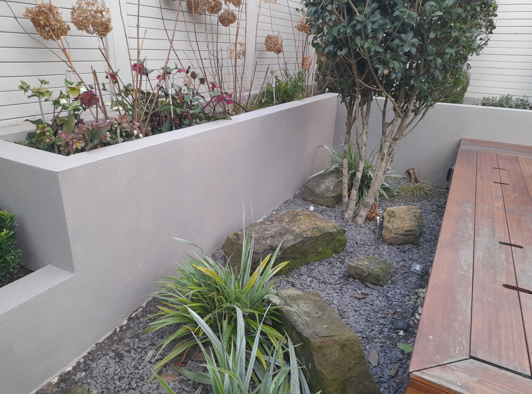 Kaskadowy ogród z roślinami w podwyższonych rabatach, kamieniami i szarym żwirem. Drewniany element architektury ogrodowej widoczny z boku.