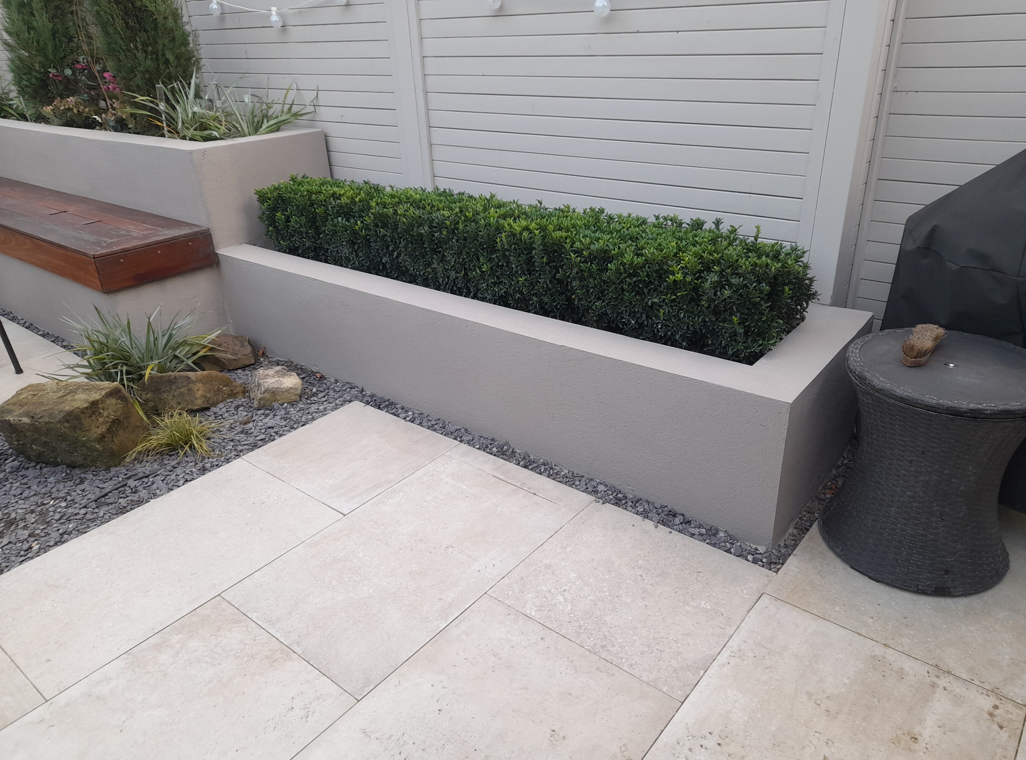 Nowoczesny ogród z podwyższonymi rabatami, drewnianą ławką i jasną kostką. Minimalistyczny design z elementami zieleni i kamienia.