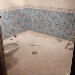 Marcin Tokarski - Łazienka w trakcie remontu: częściowo położone płytki ścienne z dekoracyjnym wzorem, przygotowane podłoże pod nowe płytki podłogowe, widoczna muszla klozetowa i drabina.