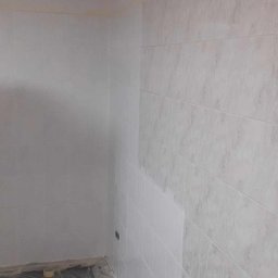 Marcin Tokarski - Wykończona ściana w łazience z białymi, marmurkowymi płytkami. Poziomica na szafce. Podłoga zabezpieczona folią. Widok na narożnik pomieszczenia.