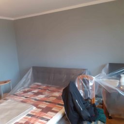 Marcin Tokarski - Pokój przed malowaniem: meble zabezpieczone folią, ściany w odcieniach szarości, listwa przysufitowa. Widok na przygotowane pomieszczenie do prac malarskich.