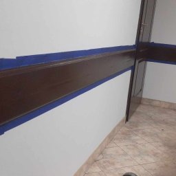 Marcin Tokarski - Świeżo pomalowana, brązowa lamperia zabezpieczona niebieską taśmą malarską na białej ścianie. Widoczna podłoga z płytek i fragment drzwi.