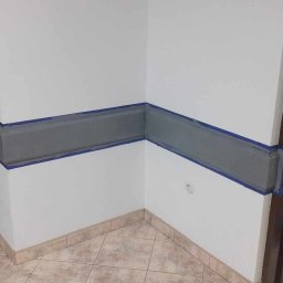 Marcin Tokarski - Przygotowanie do malowania: szara lamperia zabezpieczona taśmą malarską na białej ścianie w rogu pokoju. Widoczna podłoga z płytek i fragment otwartych, ciemnych drzwi.