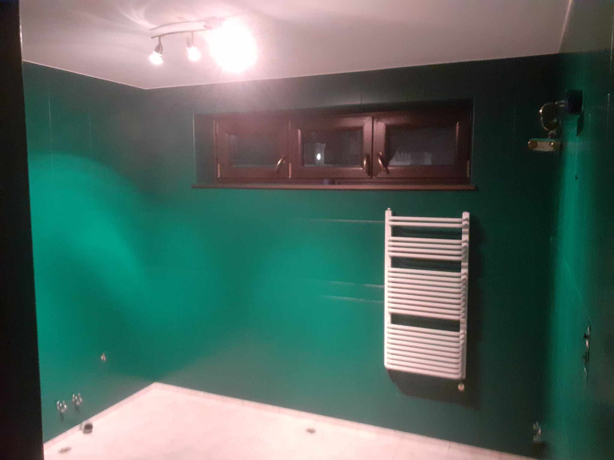 Wykończona łazienka z zielonymi ścianami, brązowym oknem i białym grzejnikiem drabinkowym. Widoczne punkty świetlne na suficie i biała podłoga.