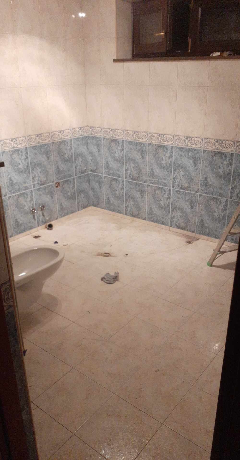 Łazienka w trakcie remontu: częściowo położone płytki ścienne z dekoracyjnym wzorem, przygotowane podłoże pod nowe płytki podłogowe, widoczna muszla klozetowa i drabina.