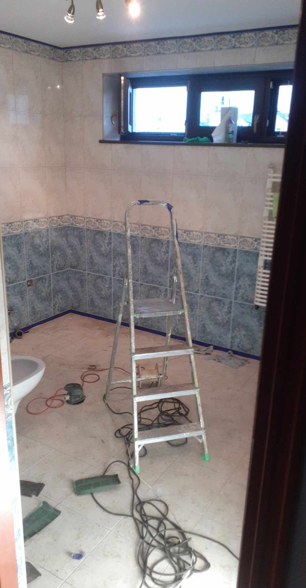 Łazienka w trakcie remontu: drabina, szlifierka i płytki z dekoracyjnym wzorem. Widoczne przygotowania do dalszych prac wykończeniowych.