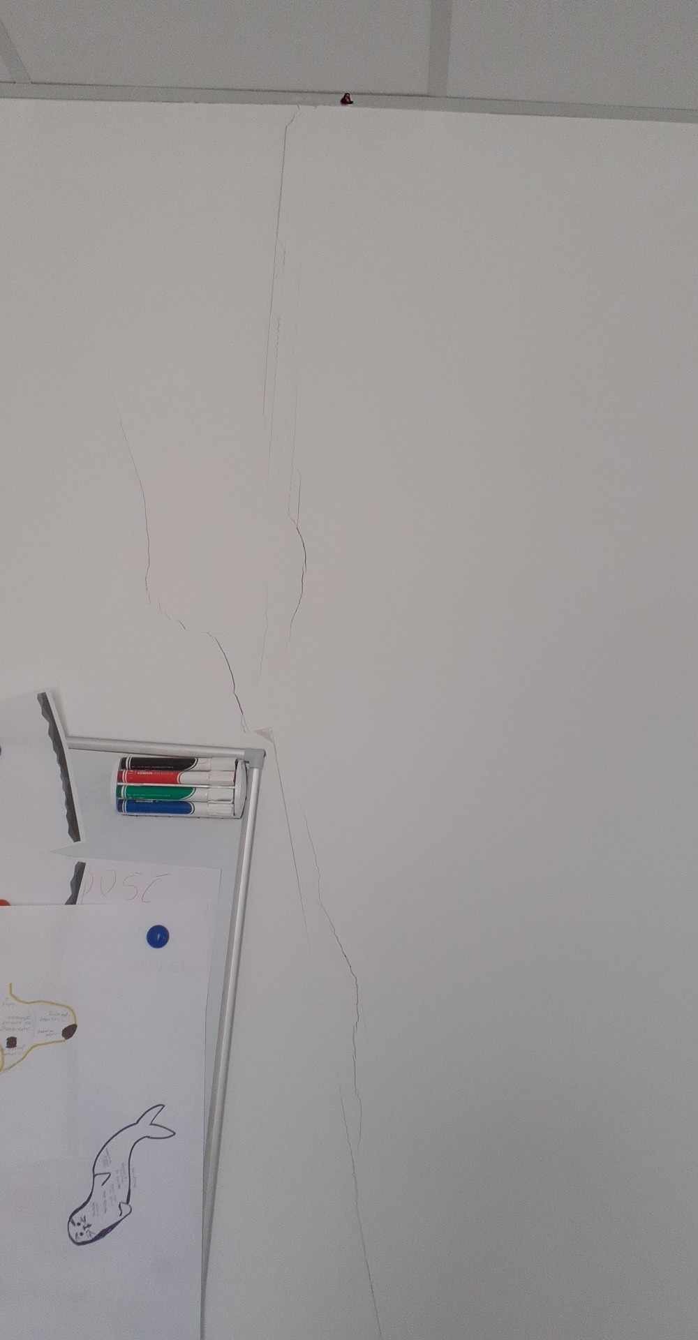 Pęknięcia na białej ścianie, widoczne obok tablicy z rysunkami i markerami. Uszkodzenia wymagają naprawy, prawdopodobnie szpachlowania i malowania.