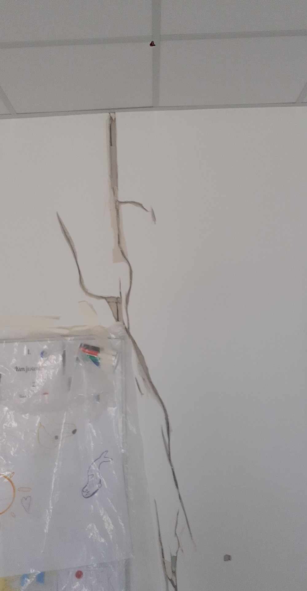 Uszkodzona ściana z widocznymi pęknięciami i ubytkami, zabezpieczona folią ochronną. Widoczne ślady po rysunkach na folii.